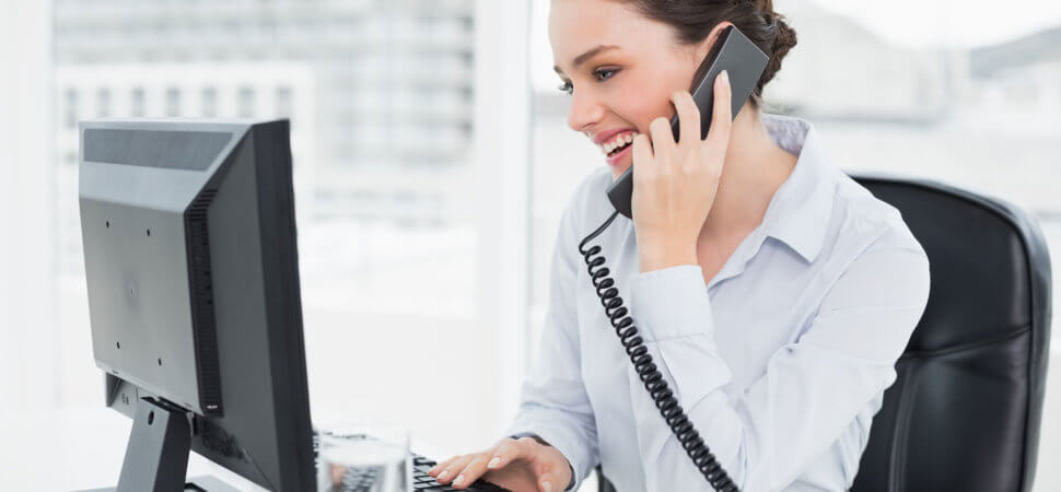 Een vrouw die aan het bellen is terwijl ze achter een computerscherm zit, glimlachend en betrokken.