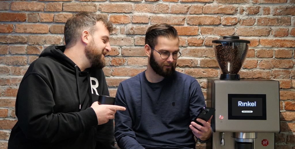 Twee mannen lachen en praten over de Rinkel App terwijl ze in een kantoor zitten, met een koffiemachine op de achtergrond.