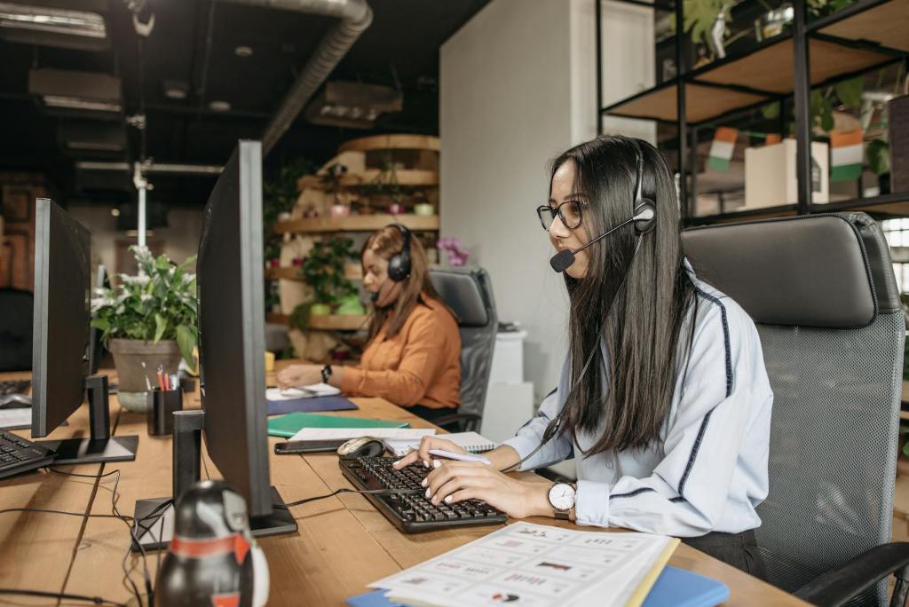 Een klantservice medewerker werkt aan een bureau met meerdere monitors en een headset, terwijl een collega in de achtergrond bezig is.