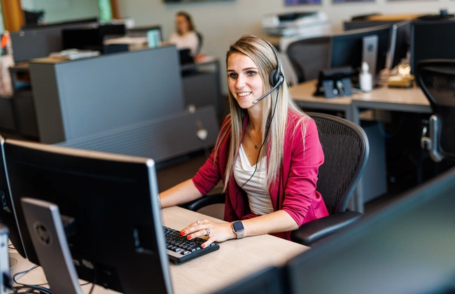 Een glimlachende vrouwelijke manager met een headset die aan het werk is achter een computerscherm in een kantooromgeving.