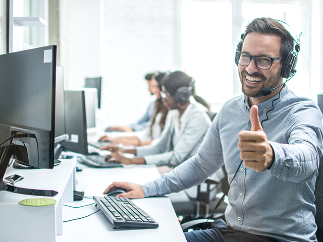 Man with glasses and headset smiling and giving a thumbs up while sitting at a desk with a computer in an office setting.